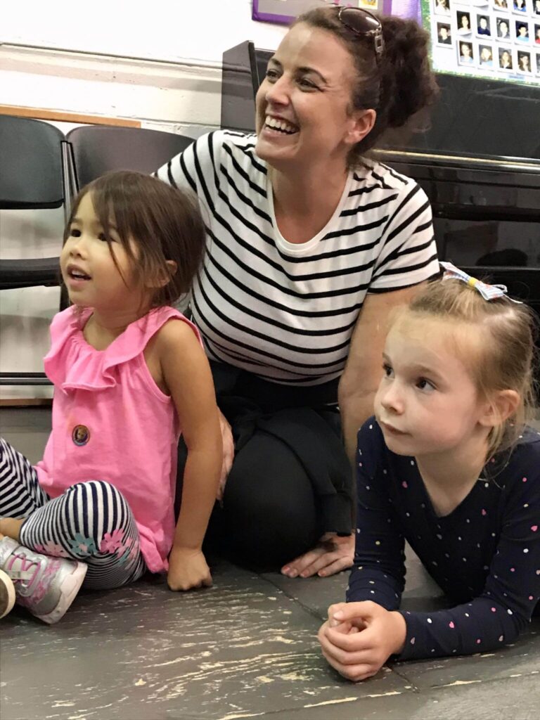 A teaching artist sits on the floor smiling with two young students during a CTA class